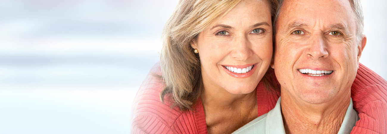The image shows a man and woman smiling at the camera  the woman has red hair and is wearing a pink top, while the man has gray hair and is dressed in a white shirt. They appear to be posing for a portrait with a blurred background.