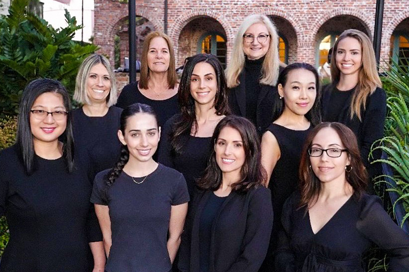 The image shows a group of people posing together outdoors, likely for a formal occasion such as a wedding or corporate event, with a woman in the center who appears to be the organizer or leader of the group.