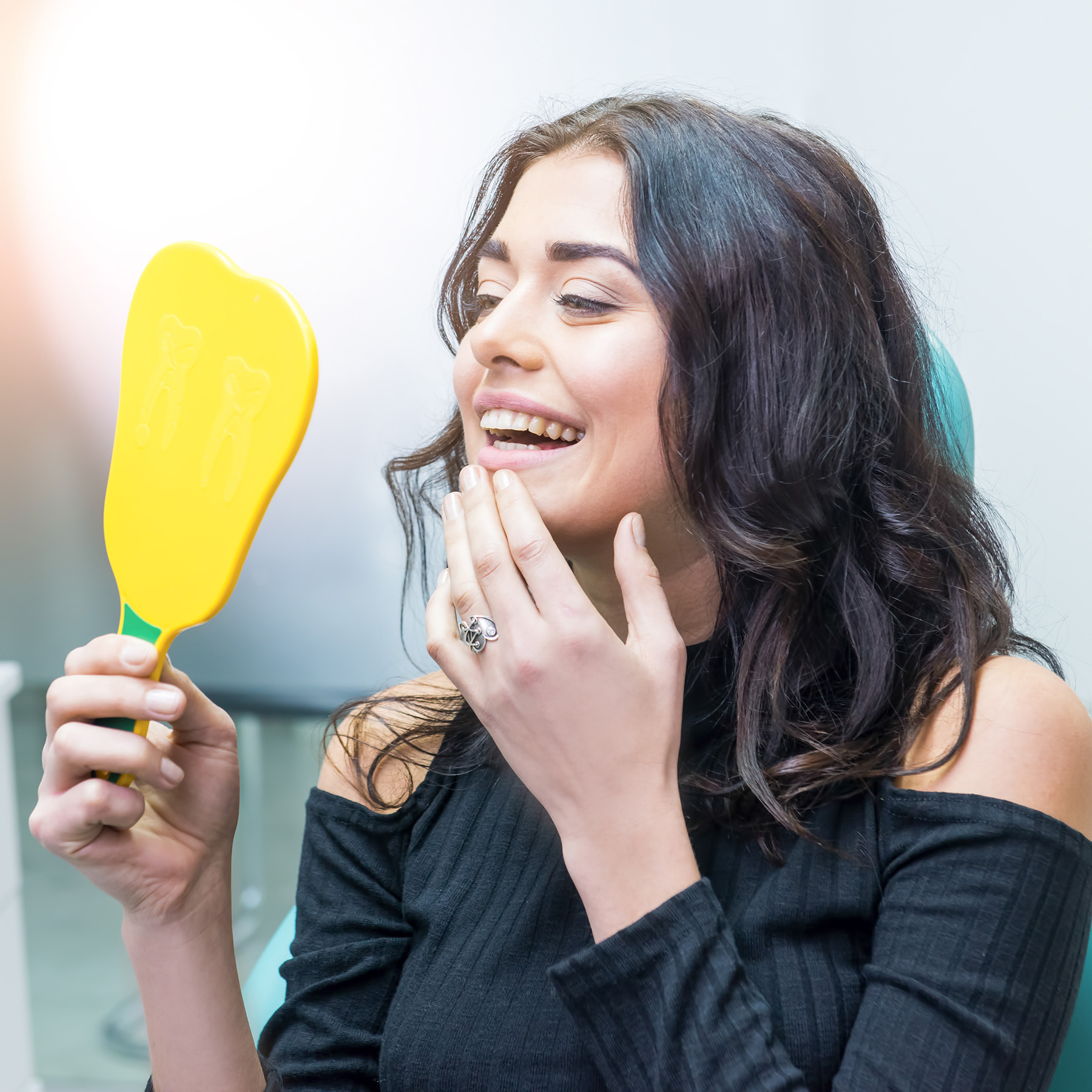 A woman with dark hair holding a yellow makeup brush in front of her face, smiling while looking at her reflection in a mirror.
