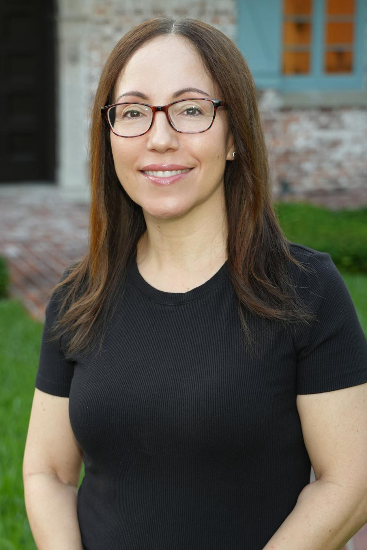 The image shows a woman standing outdoors, wearing glasses, a black top, and smiling at the camera. She has long hair and appears to be posing for the photograph.