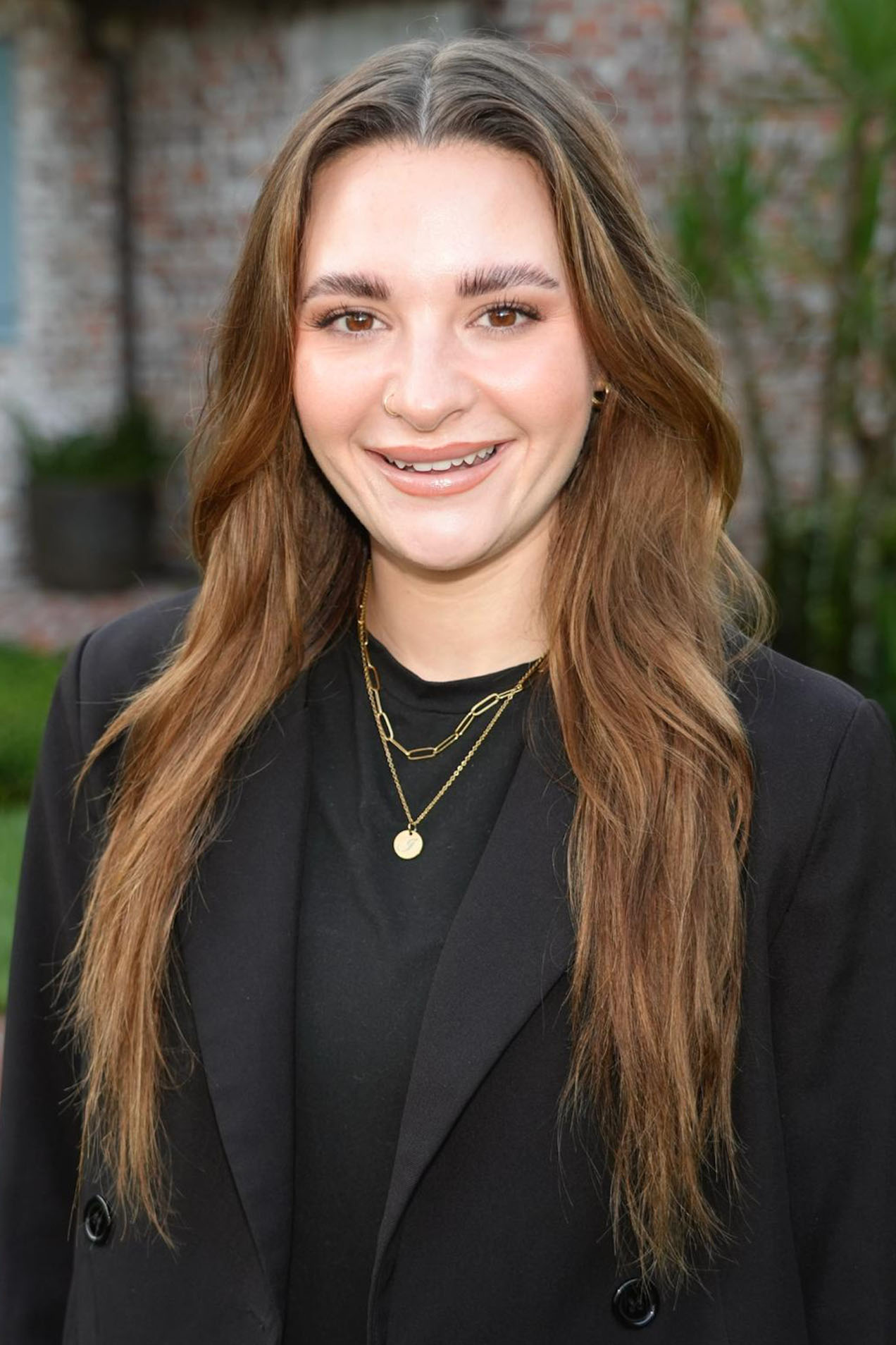 The image shows a young woman with long hair, smiling slightly, wearing a black blazer and a necklace, standing outdoors with a building in the background.
