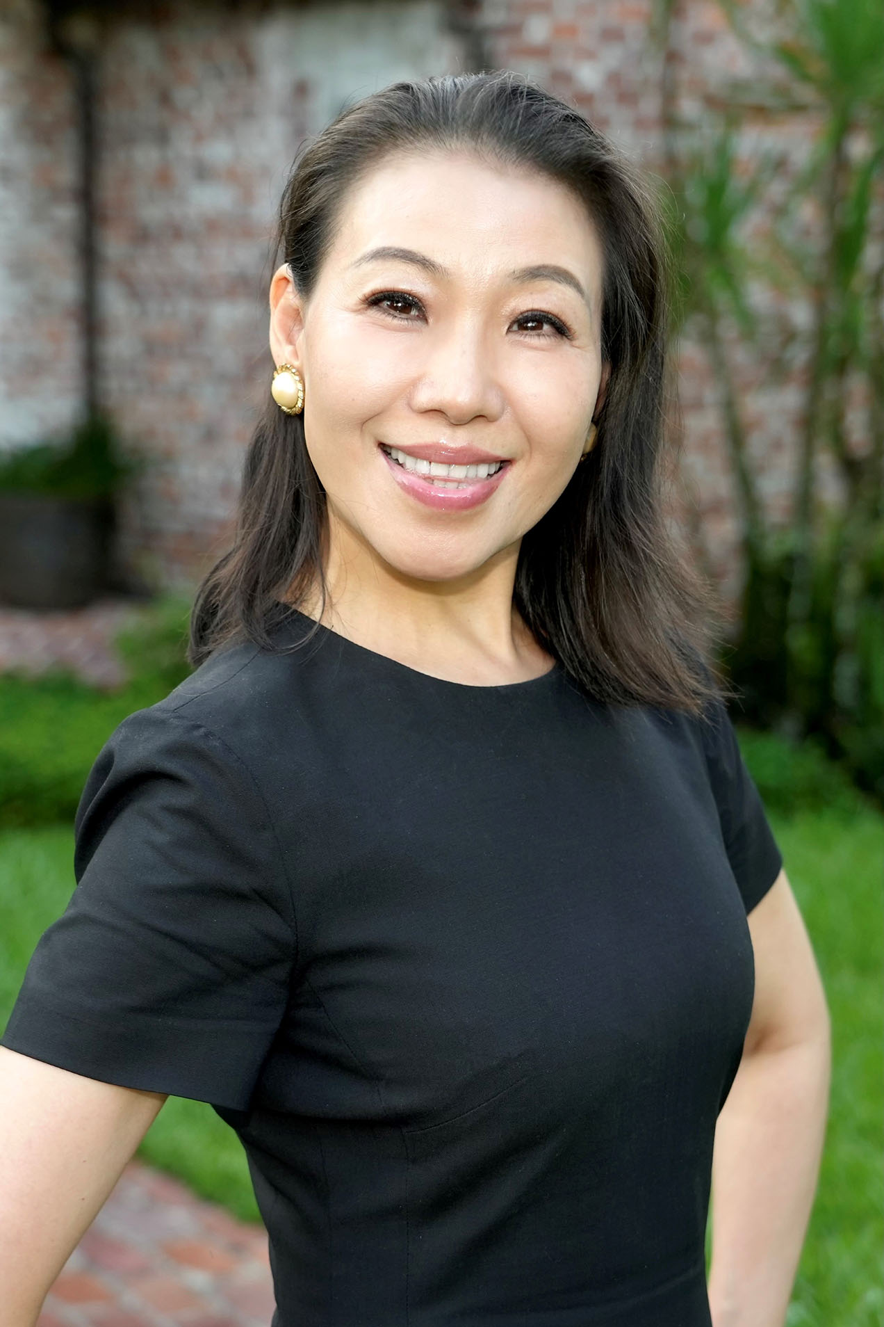The image shows a woman with short hair smiling at the camera while wearing a black top. She stands outdoors against a backdrop of greenery and a building.